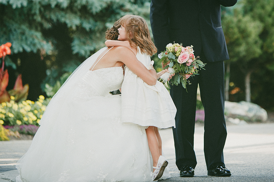A flower-girl stands on tippy-toes to hug the bride.