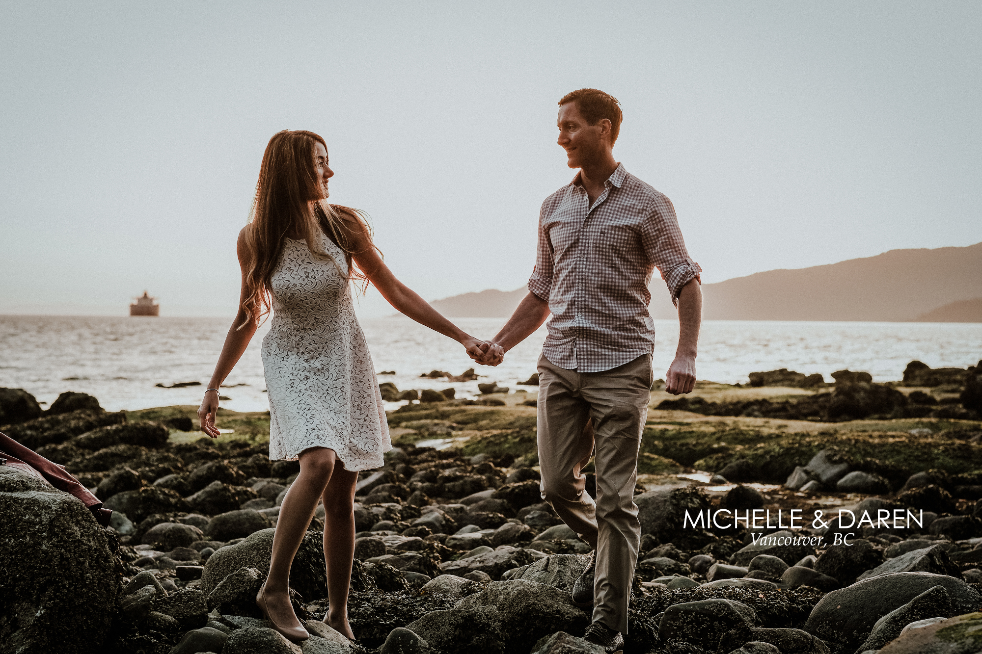 Man and woman walk precariously on rocky beach during engagement session.
