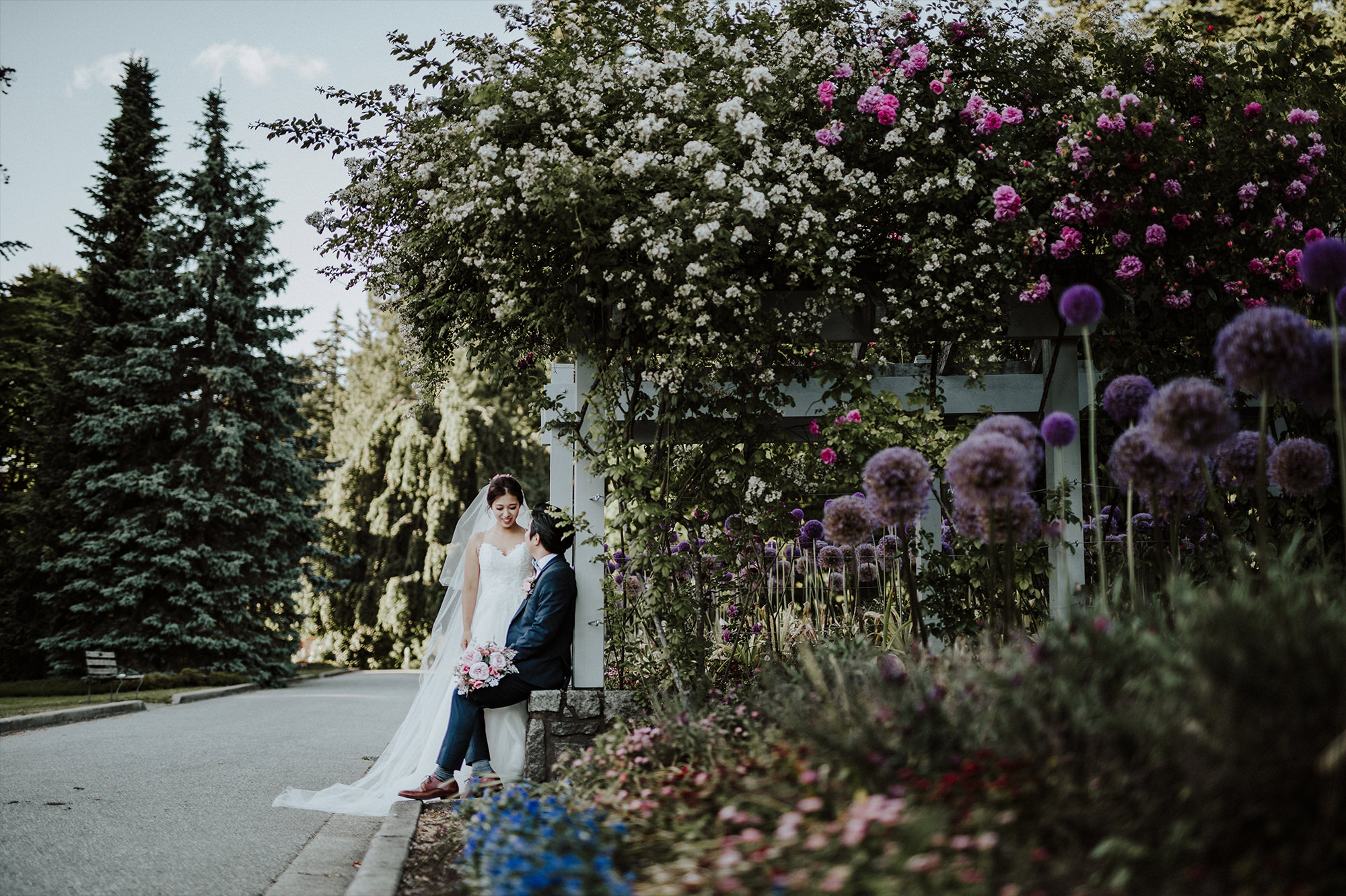 Seated groom invites his Korean bride to sit on his lap in a garden